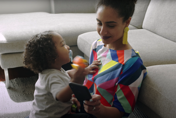 woman in a rainbow geometric shirt and yellow earrings looks at her phone with toddler son on her lap