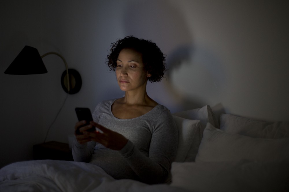 woman sits in dark room in bed and looks at phone