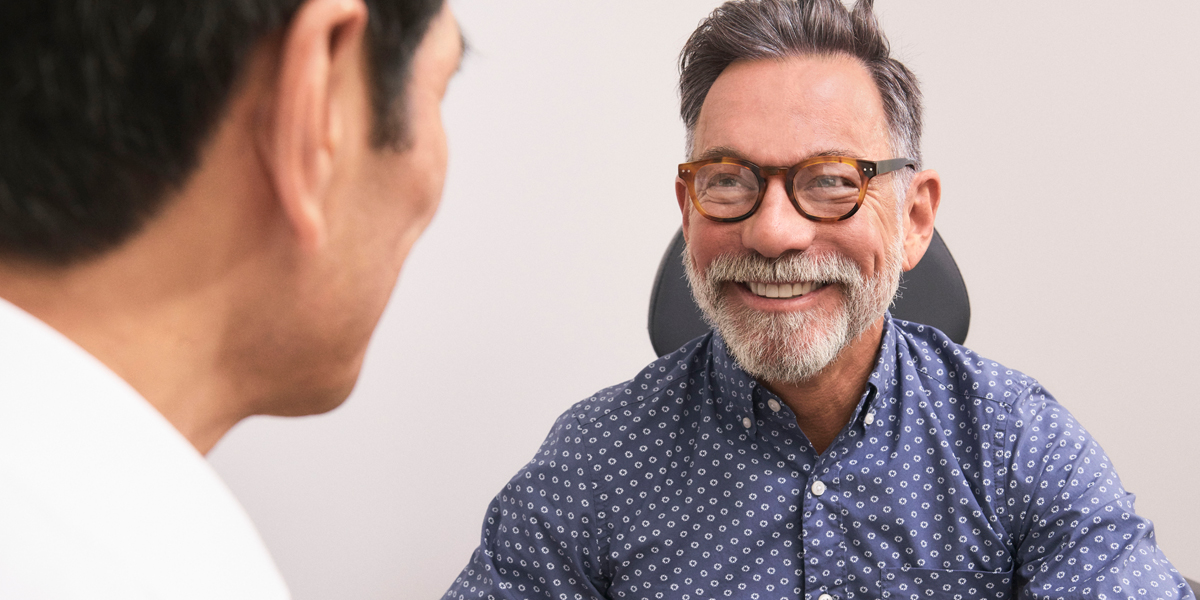 man in round glasses and polka dot button up smiles at another man