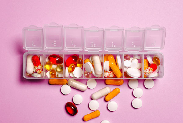large group of vitamins and supplements sitting in and around a weekly pill organizer on a pink background