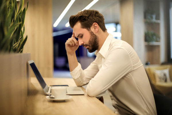 stressed man sits at desk in front of laptop with head in hand